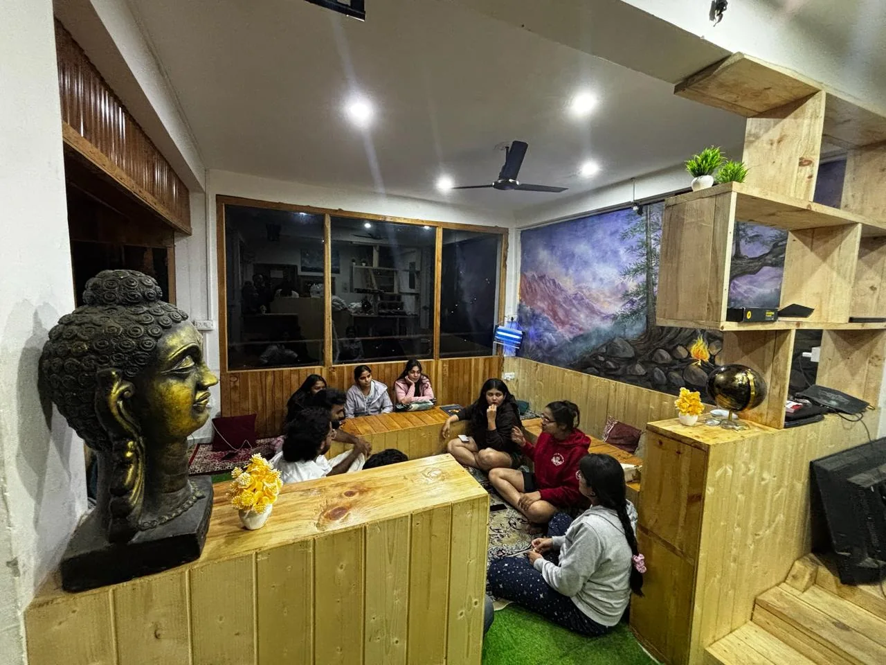 Guests relaxing around wooden tables in the café, with a bronze Buddha head and a painted mountain mural