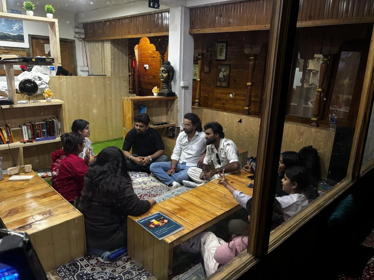 Guests chatting on low wooden tables in the Jibhi Emberwood café, seen through a wooden-framed window