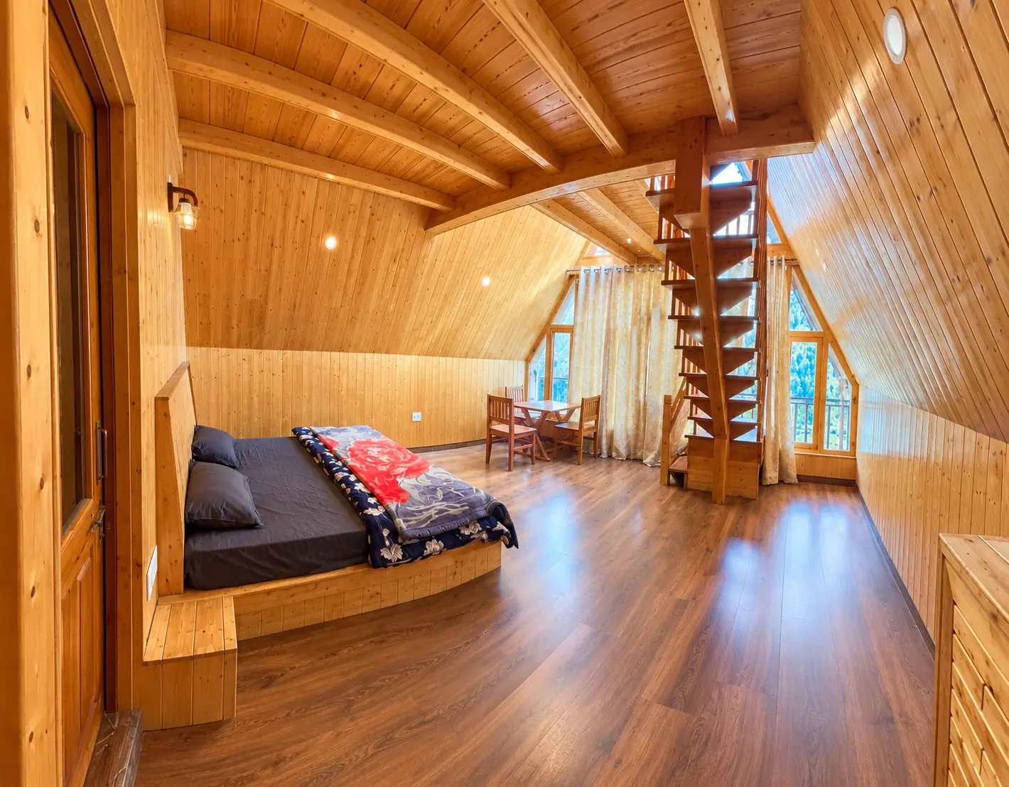 Spacious loft bedroom with pine-panelled walls and a sloping ceiling, a large bed, a small dining table with chairs and a wooden staircase beside tall windows