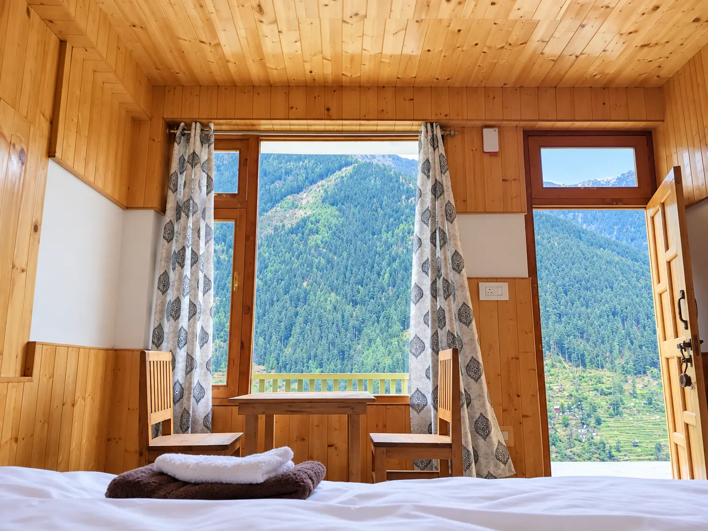 Pine-panelled bedroom seen from the bed, with folded towels, two chairs and a table in front of a large window and an open door looking onto forested mountains
